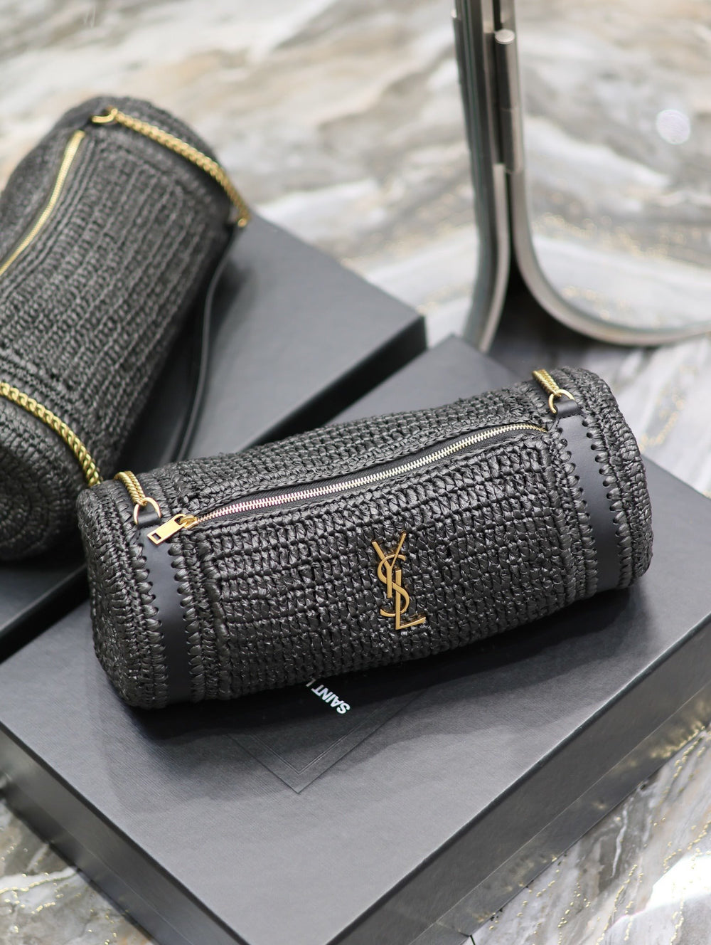 Natural raffia cylindrical bag with bronze chain strap coiled on dark marble surface
