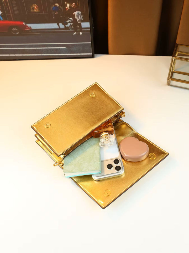 A gold metallic zippered clutch wallet shown open on a white tabletop, filled with a light blue cardholder, a smartphone, a clear case, and a pink heart-shaped compact.