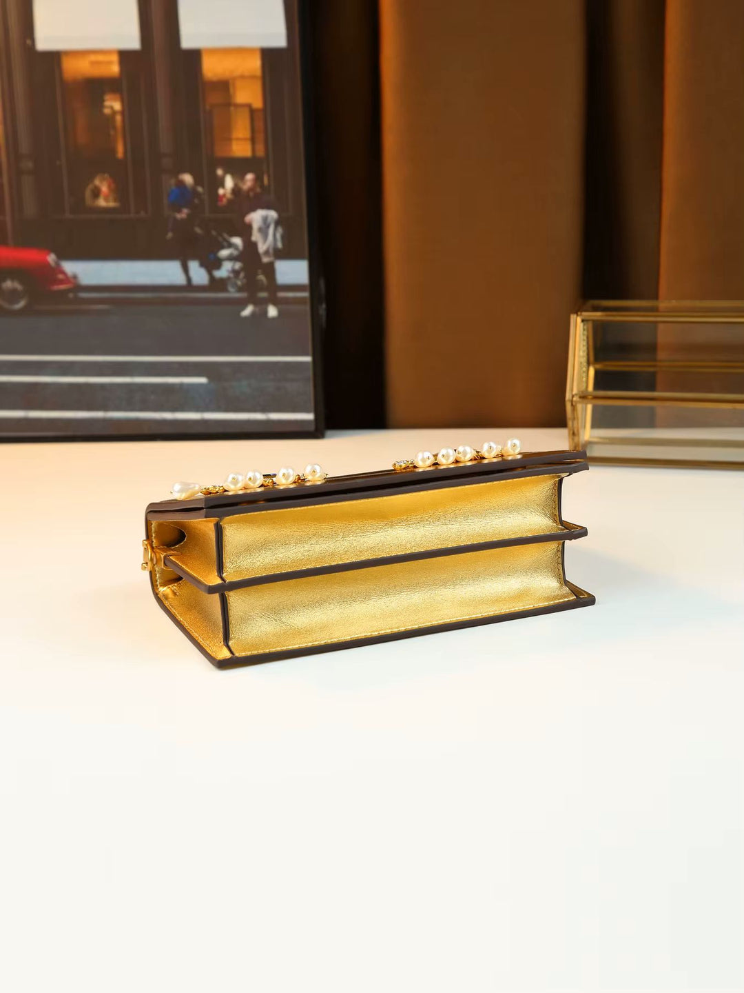 A small book-shaped clutch purse with a textured gold interior, black faux-leather edges, and a row of white pearls along the top, displayed on a white surface.