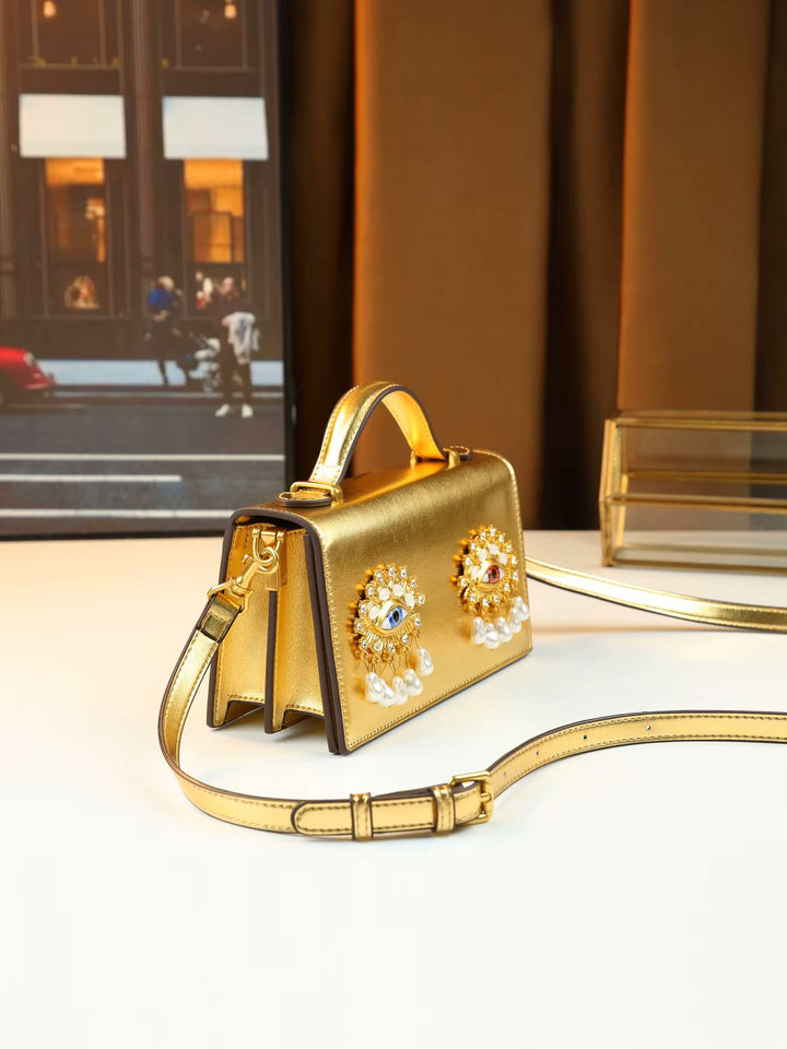 A gold metallic mini handbag with a top handle and shoulder strap, decorated with two large floral brooch embellishments featuring pearls and colorful rhinestones, displayed on a white surface.