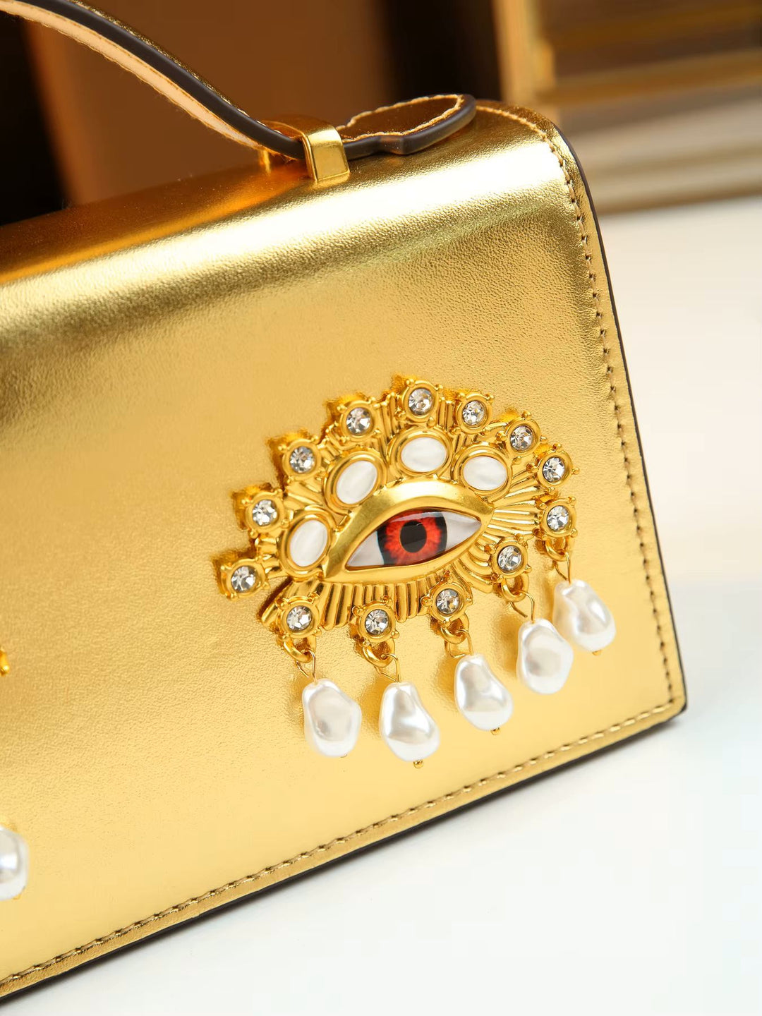Close-up of a metallic gold handbag featuring an ornate gold evil eye brooch embellished with clear rhinestones, white pearl-like drops, and a red iris detail.