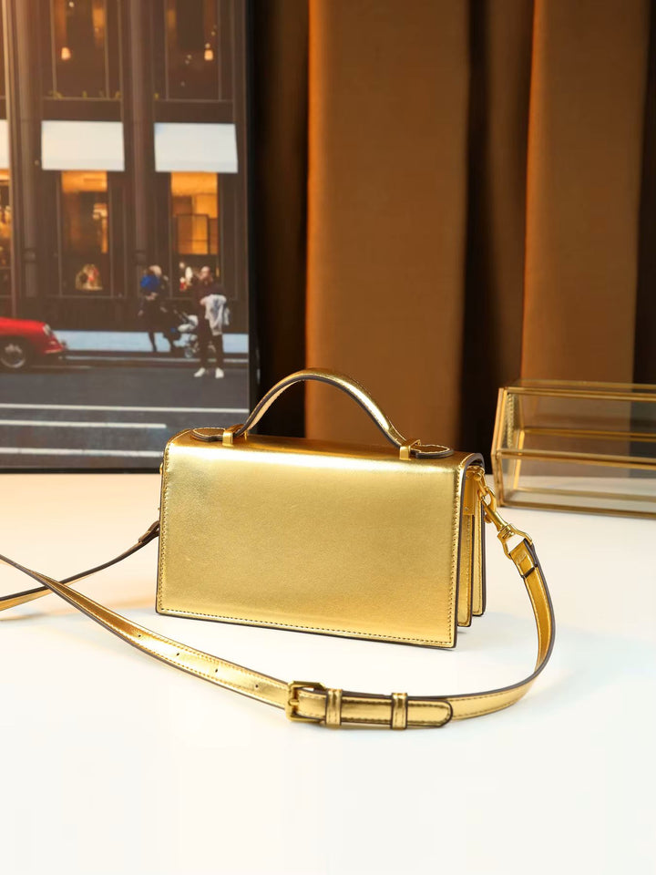 A metallic gold structured handbag with a top handle and matching shoulder strap, displayed on a white surface with a warm brown curtain and framed city scene in the background.