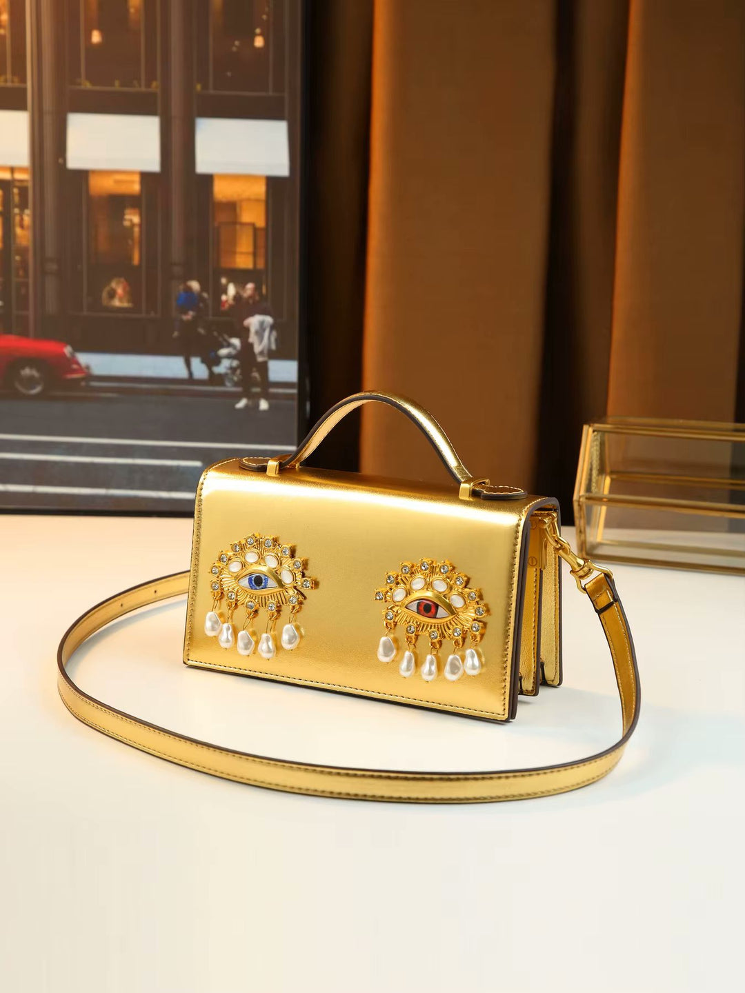 A metallic gold handbag with a top handle and shoulder strap, decorated with two ornate jeweled eye motifs on the front, displayed on a tabletop with a warm indoor background.