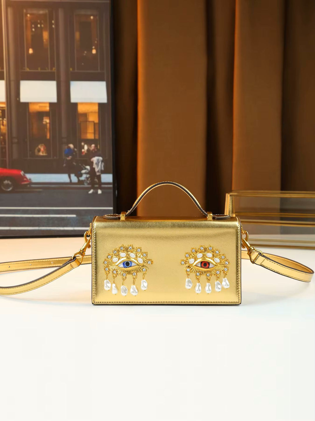 A gold metallic handbag with a top handle and shoulder strap, featuring two ornate evil eye brooch embellishments on the front. The bag is displayed on a white surface with a warm brown curtain and a framed picture in the background.