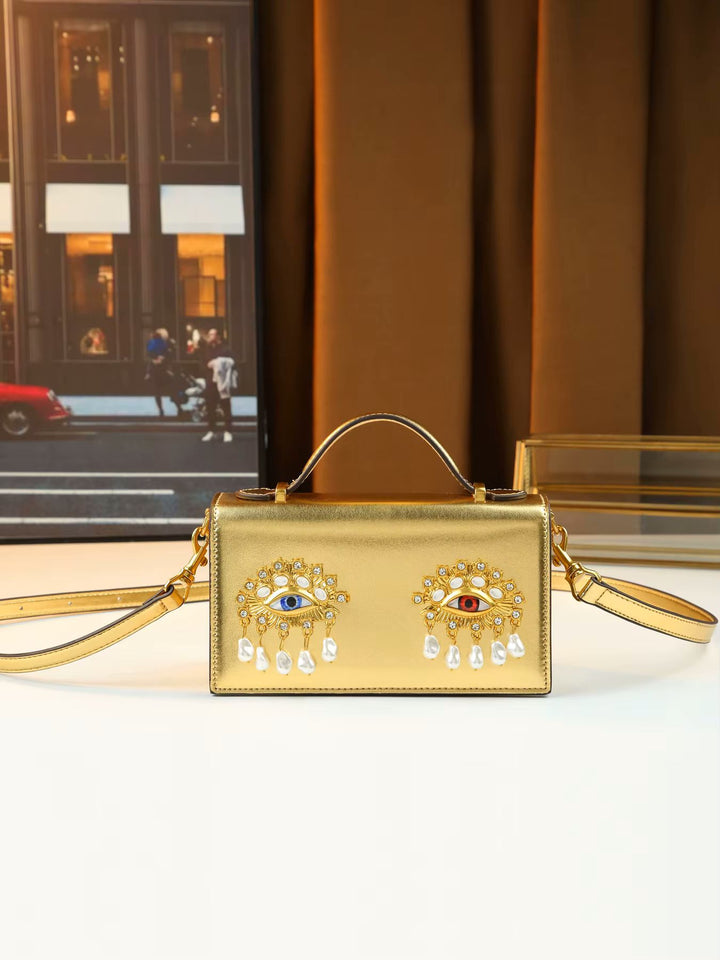 A gold metallic handbag with a top handle and shoulder strap, featuring two ornate evil eye brooch embellishments on the front. The bag is displayed on a white surface with a warm brown curtain and a framed picture in the background.