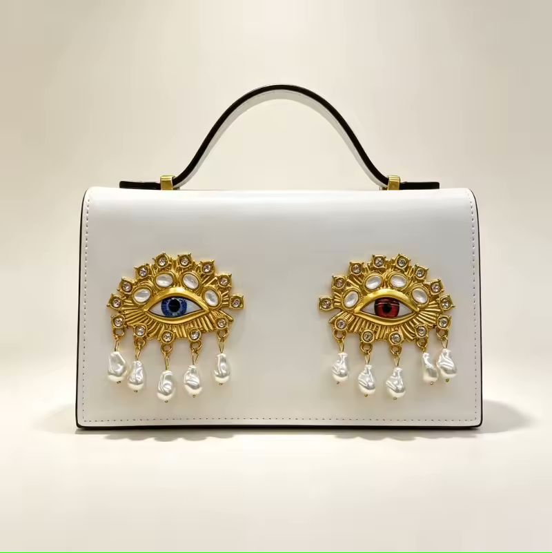 Gold leather small square bag with 3D eye embellishment lifestyle shot at styled flat lay