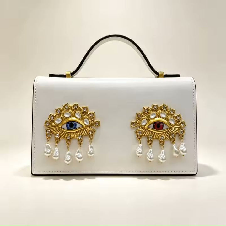 Gold leather small square bag with 3D eye embellishment lifestyle shot at styled flat lay