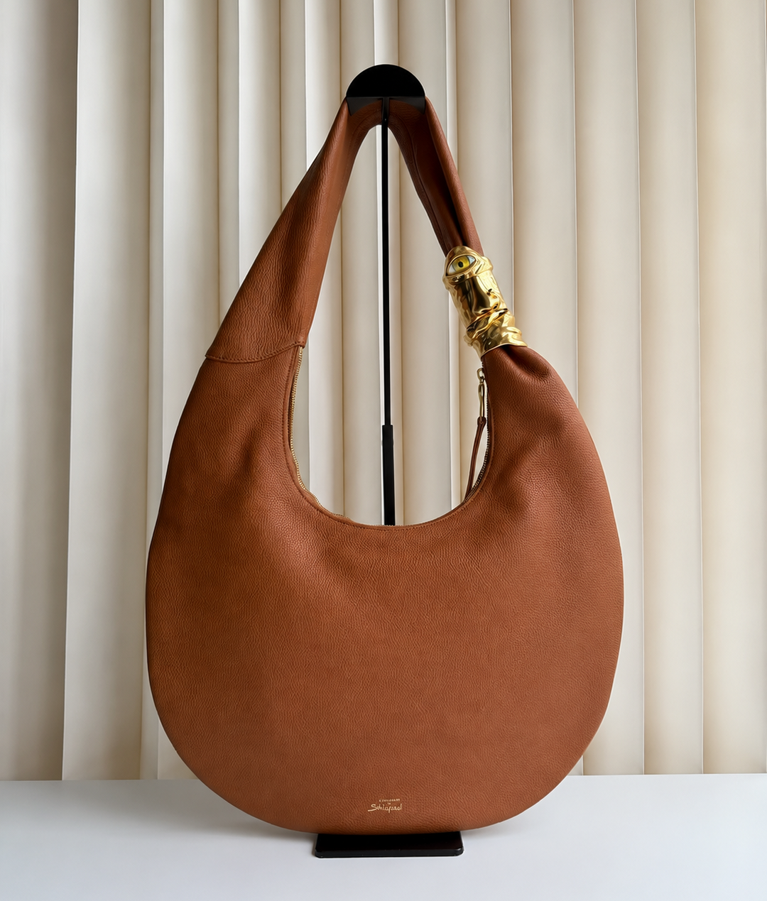 Brown leather handbag with gold clasp on a stand against a neutral background