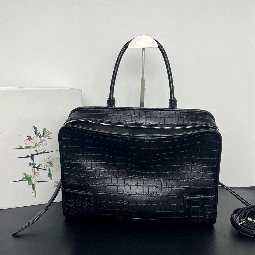 A black crocodile-embossed leather handbag with double top handles, displayed on a dark surface against a light gray background.