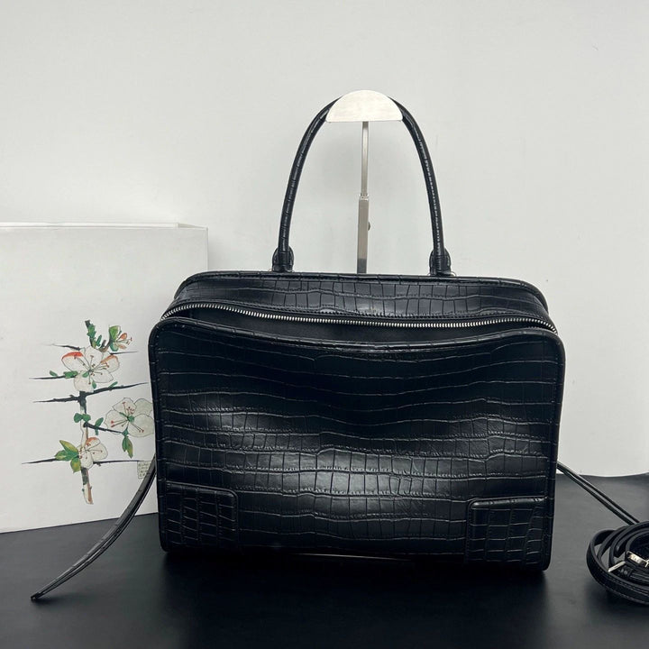 A black crocodile-embossed leather handbag with double top handles, displayed on a dark surface against a light gray background.
