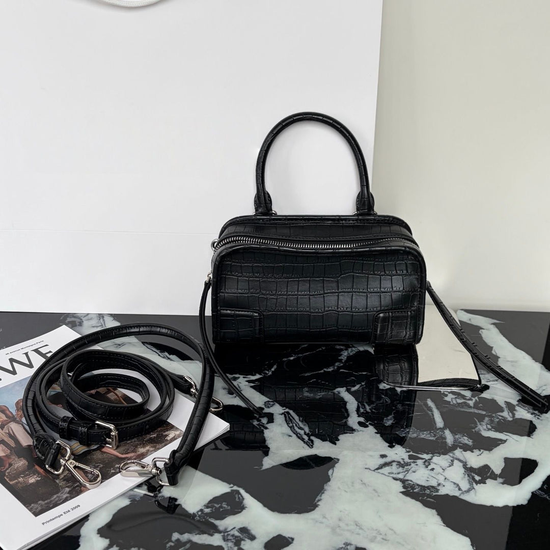 Black crocodile-embossed leather handbag with top handles and a detachable shoulder strap, displayed on a black-and-white marble surface with a magazine nearby.