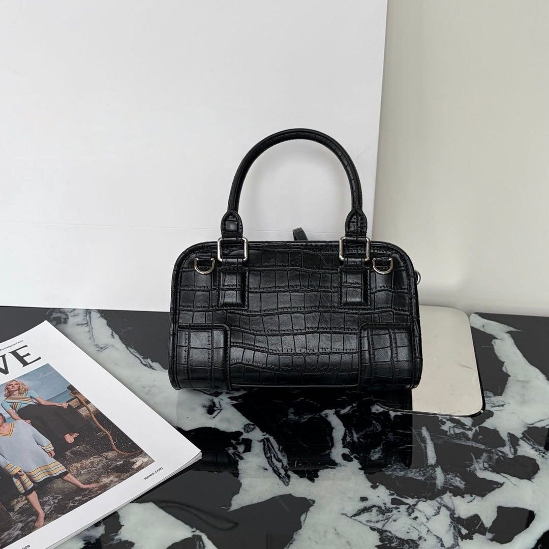 A structured black crocodile-embossed leather handbag with double top handles and silver-tone hardware, displayed on a black-and-white marble surface next to a magazine.