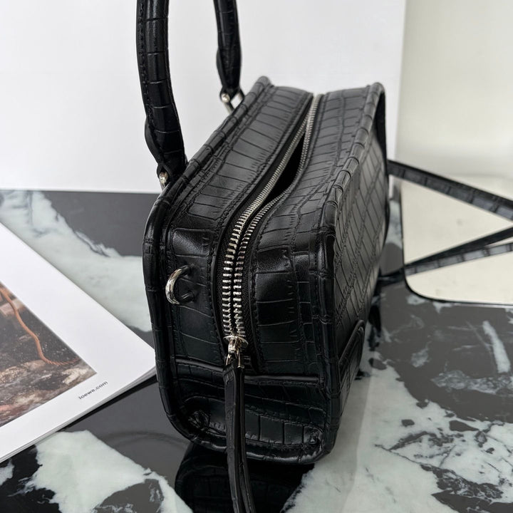 A black crocodile-embossed leather handbag with silver-tone hardware, shown on a black and white marble surface next to a mirror and printed page.