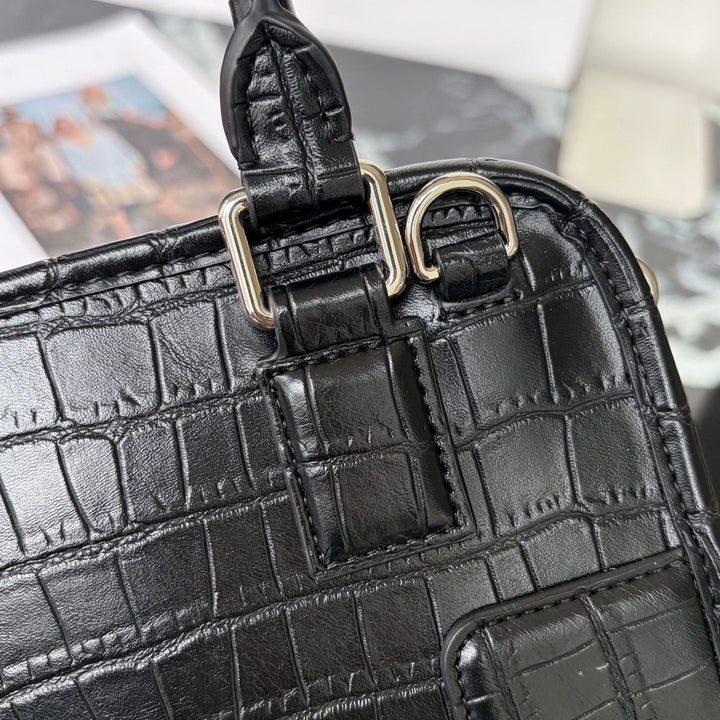 Close-up of a black crocodile-embossed leather handbag showing a strap attachment with gold-tone metal rings and hardware.