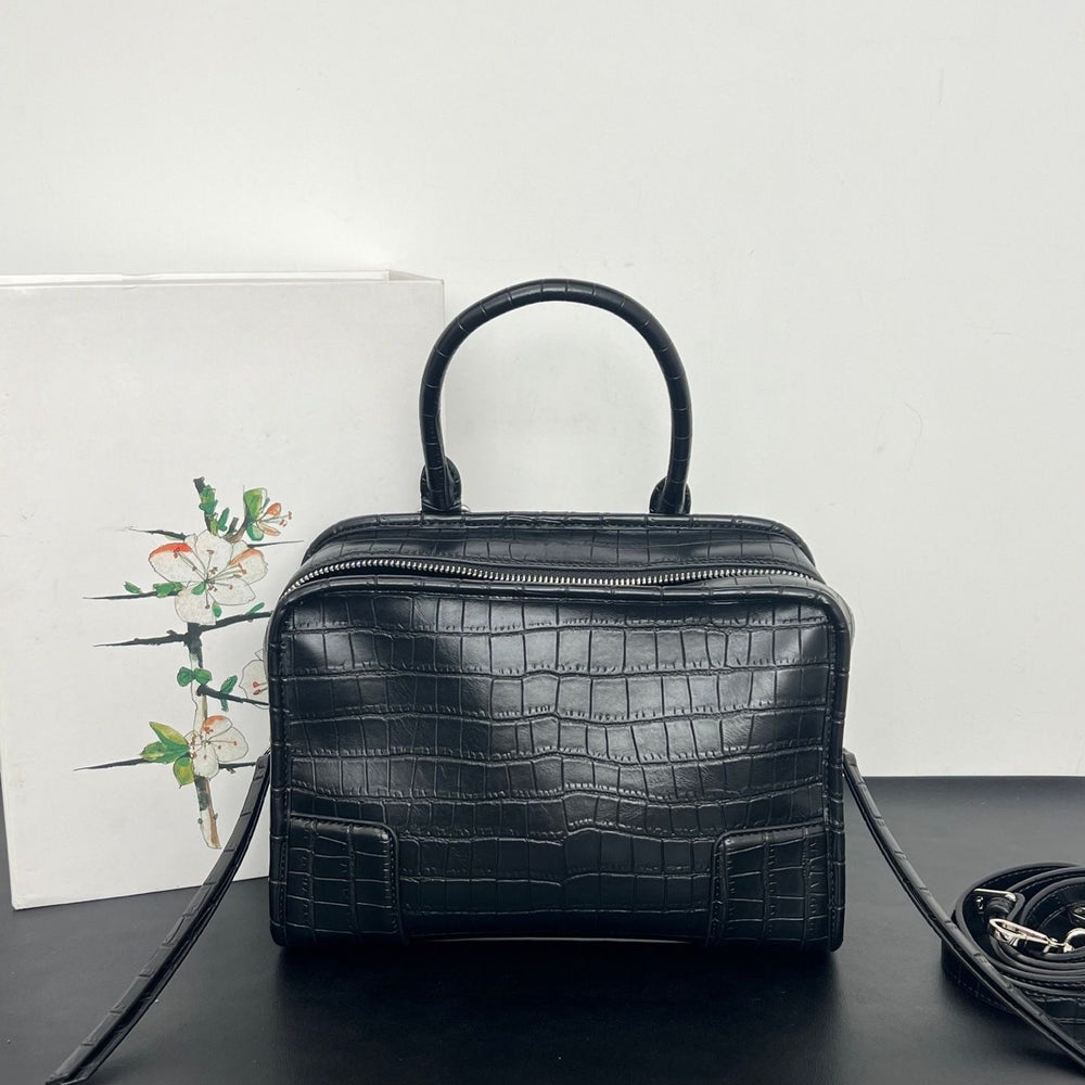 Black crocodile-embossed leather handbag with a top handle, shown on a dark surface against a white wall, with a floral card and a coiled strap nearby.