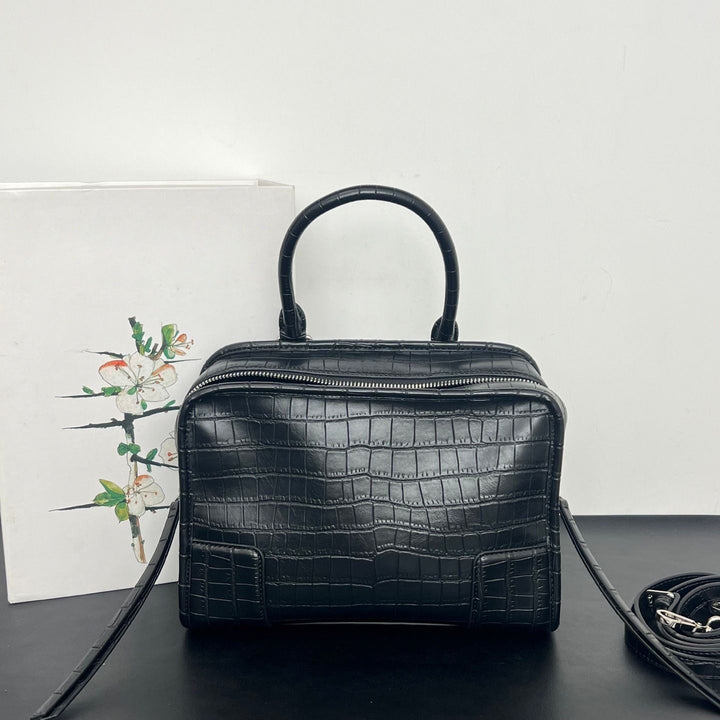 Black crocodile-embossed leather handbag with a top handle, shown on a dark surface against a white wall, with a floral card and a coiled strap nearby.