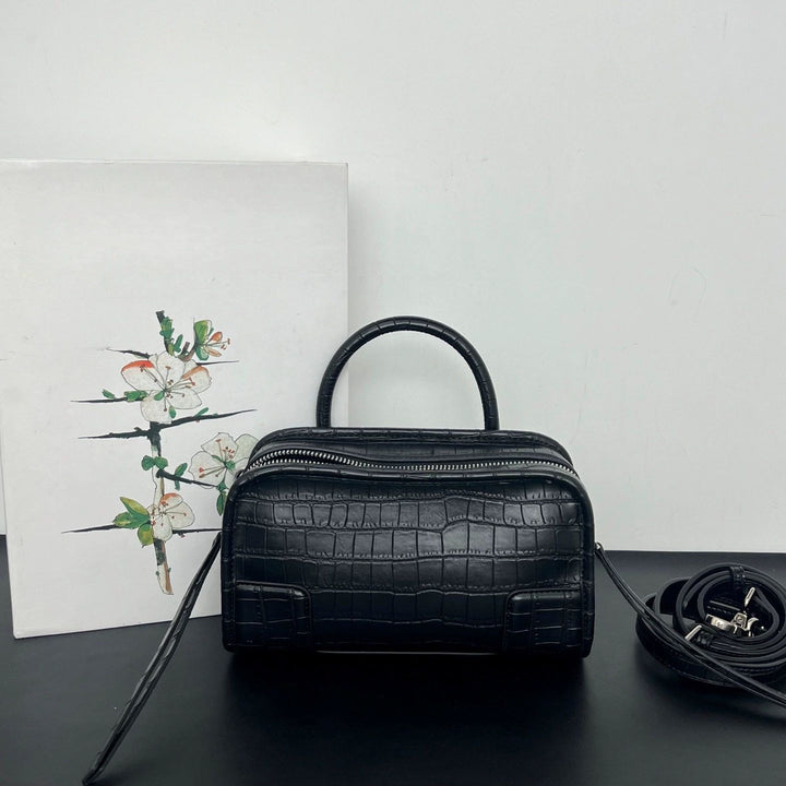 A black crocodile-embossed leather handbag with a top handle and a zippered top, displayed on a black surface against a white wall, with a floral print card and a coiled strap beside it.