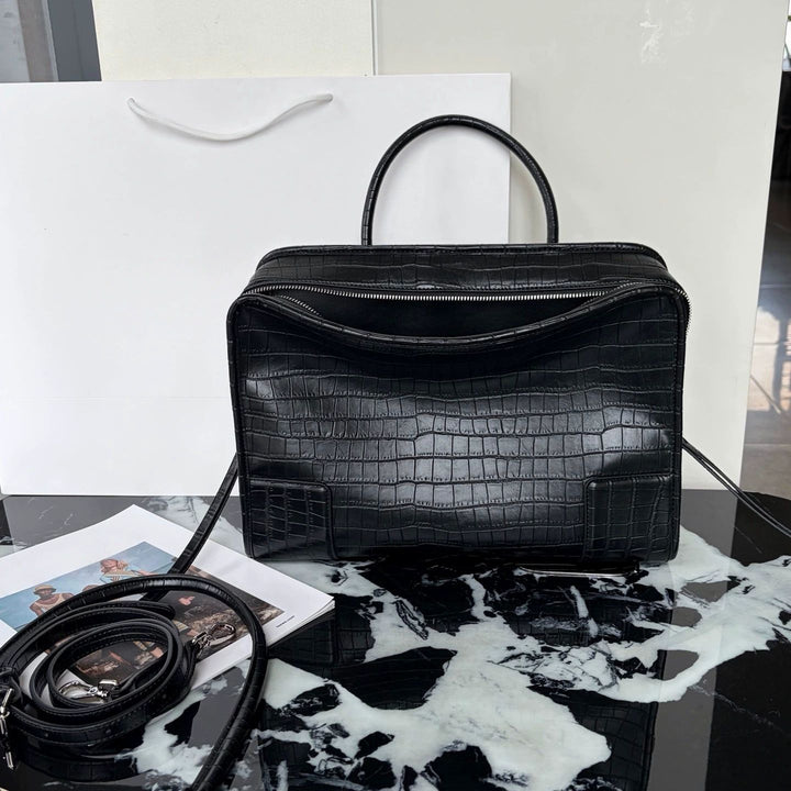 A black crocodile-embossed leather handbag with a top handle, displayed on a black and white marble surface next to a white shopping bag and a coiled black strap.