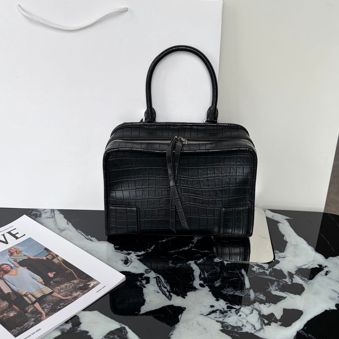 A structured black crocodile-embossed handbag with dual top handles, displayed on a black-and-white marble surface against a white wall, with a magazine partially visible in the lower left.