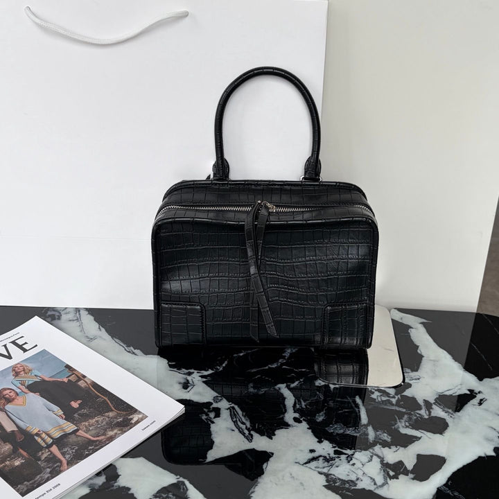 A structured black crocodile-embossed handbag with dual top handles, displayed on a black-and-white marble surface against a white wall, with a magazine partially visible in the lower left.