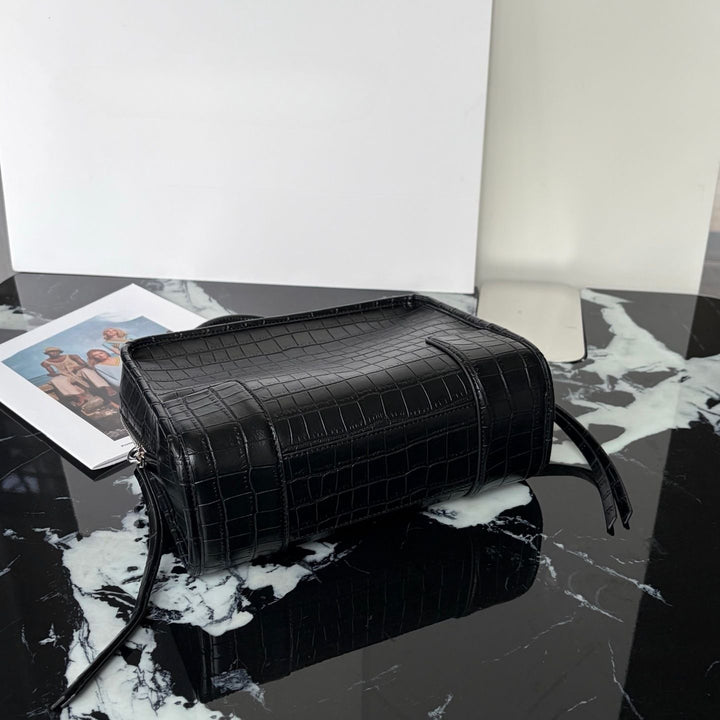 A black crocodile-embossed leather crossbody bag with a zippered top and long strap, resting on a black and white marble surface.