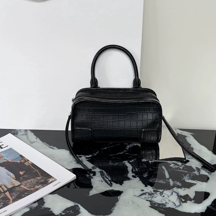 A black crocodile-embossed leather handbag with a top handle and shoulder strap, displayed on a black and white marble surface next to an open magazine.
