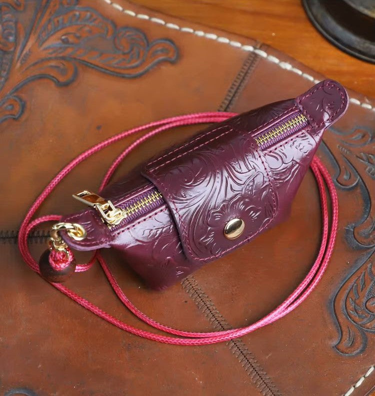 A small embossed leather pouch in deep burgundy with gold-tone hardware and a matching pink braided strap, photographed on a brown tooled leather surface.