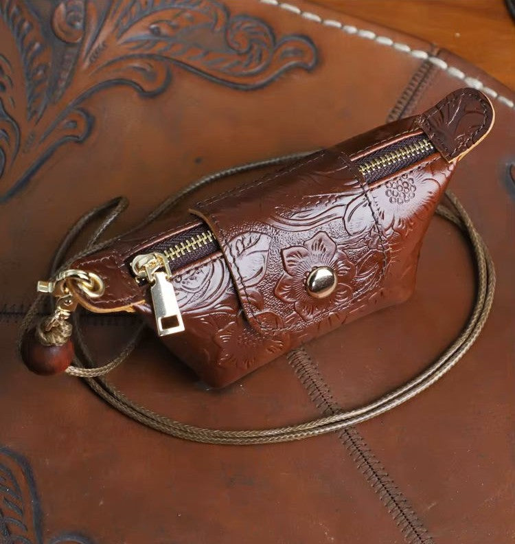 A brown embossed leather pouch with floral tooling, gold-tone zippers, and a matching leather strap, displayed on a brown leather surface with carved detailing.