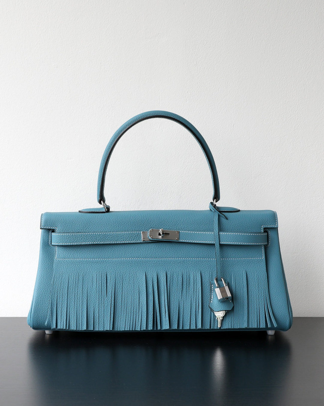 A structured turquoise blue leather handbag with a top handle, silver-tone hardware, and a long fringe detail across the front, displayed on a dark reflective surface against a plain light wall.