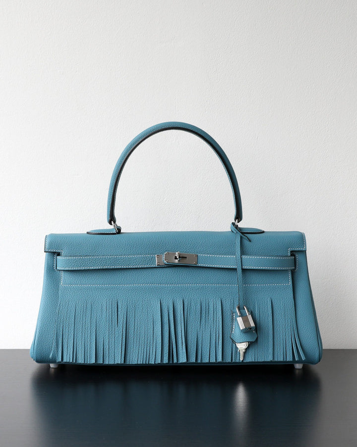 A structured turquoise blue leather handbag with a top handle, silver-tone hardware, and a long fringe detail across the front, displayed on a dark reflective surface against a plain light wall.