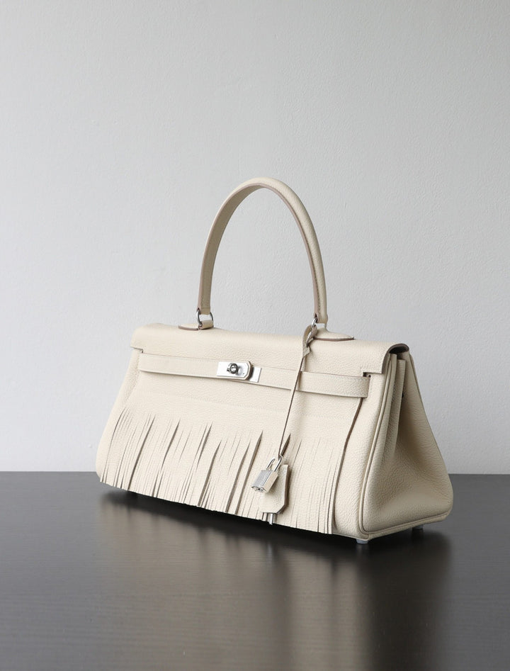 A structured cream-colored leather handbag with a distinctive fringe detail along the bottom front, a top handle, and silver-tone hardware, resting on a dark reflective surface against a light gray background.
