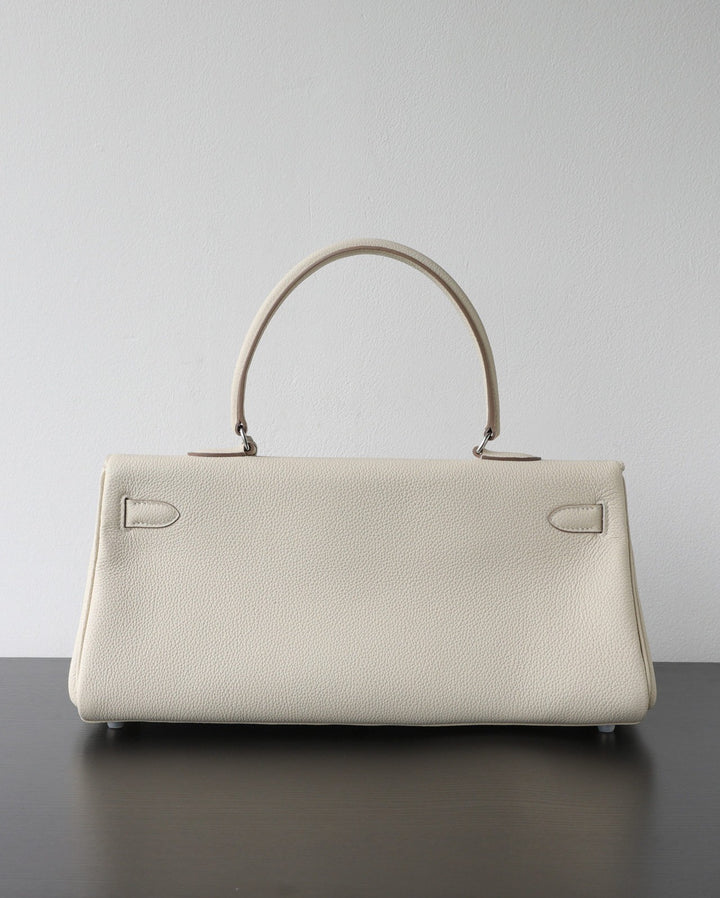 A structured cream-colored pebbled leather handbag with a top handle, resting on a dark surface against a plain light gray wall.