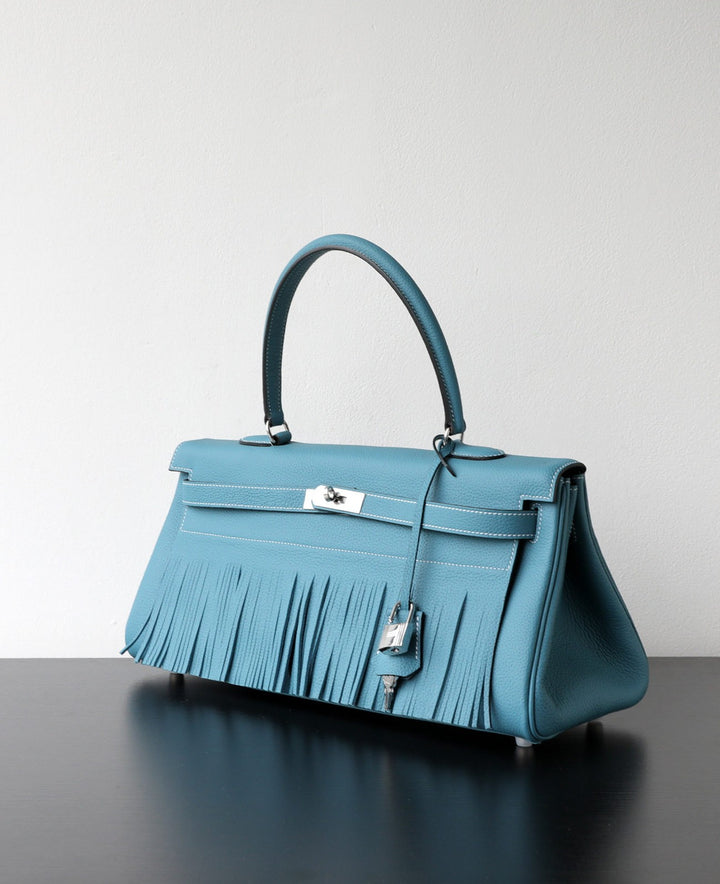 A structured blue leather handbag with a long fringe detail across the front, a top handle, and silver-tone hardware, displayed on a dark reflective surface against a light gray background.