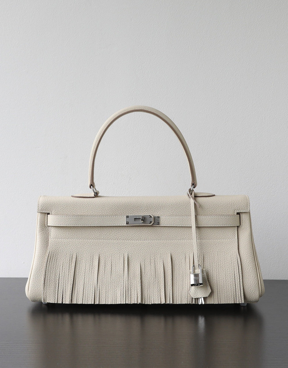 A structured cream-colored leather handbag with a fringed front, a single top handle, and silver-tone hardware, displayed on a dark surface against a light gray background.