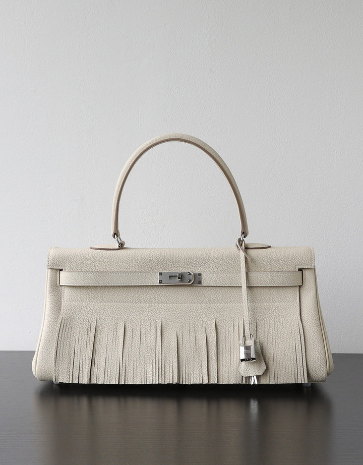 A structured cream-colored leather handbag with a fringed front, a single top handle, and silver-tone hardware, displayed on a dark surface against a light gray background.