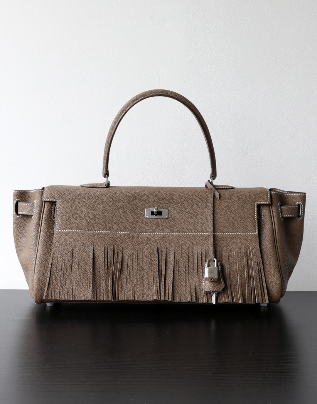 A taupe leather handbag with a fringed front detail, dual top handles, and silver-tone hardware, displayed on a dark surface against a light gray background.