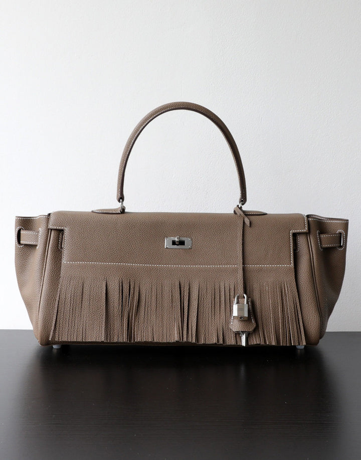 A taupe leather handbag with a fringed front detail, dual top handles, and silver-tone hardware, displayed on a dark surface against a light gray background.
