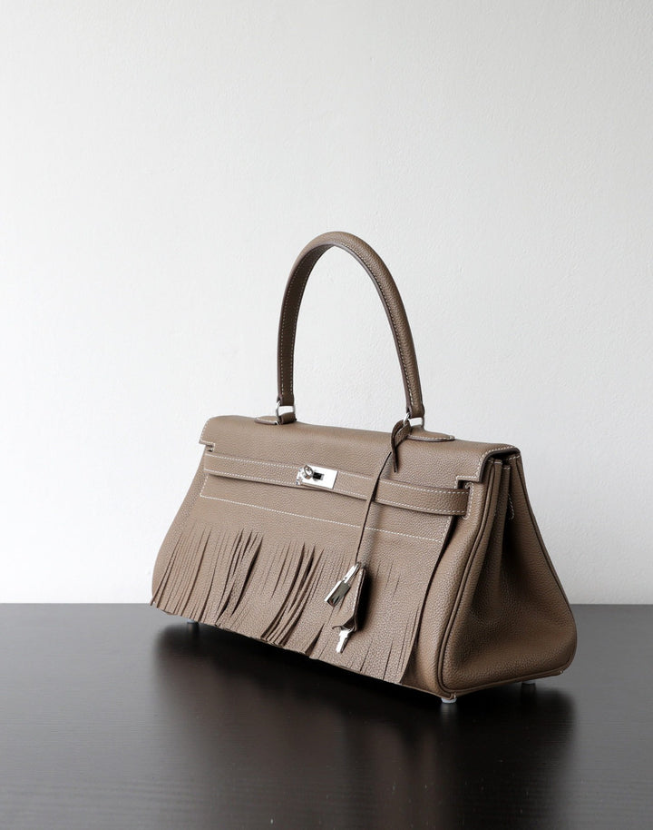 A taupe leather handbag with fringe detailing and silver-tone hardware, displayed on a dark reflective surface against a light gray background.
