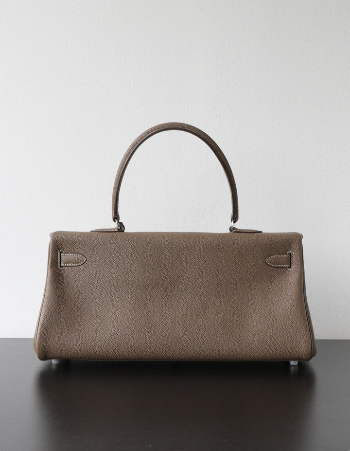 A structured taupe leather handbag with a single top handle, resting on a dark surface against a plain light gray wall.
