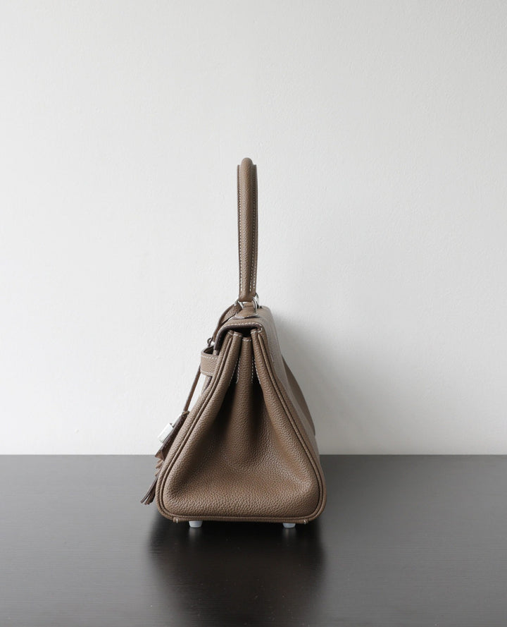 A taupe pebbled leather handbag shown from the side profile, resting on a dark surface against a plain light gray wall.