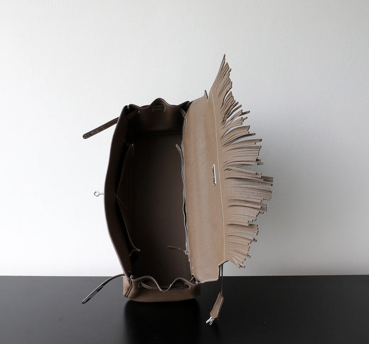 A taupe leather bucket bag with a prominent fringed flap, shown open to display its interior. The bag is positioned on a black surface against a plain white wall.