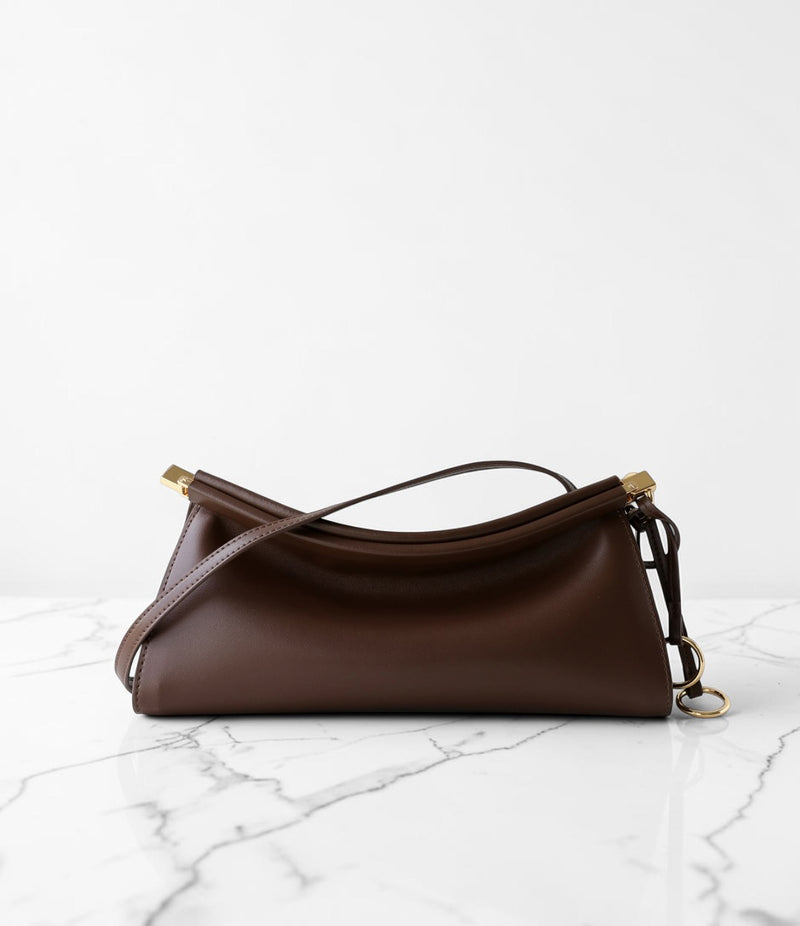 A dark brown leather shoulder bag with gold-tone hardware, resting on a white marble surface against a white background.