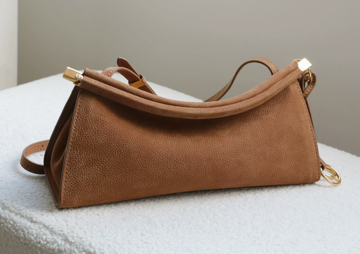 A medium brown textured leather shoulder bag with gold-tone hardware, resting on a white textured surface against a neutral gray background.