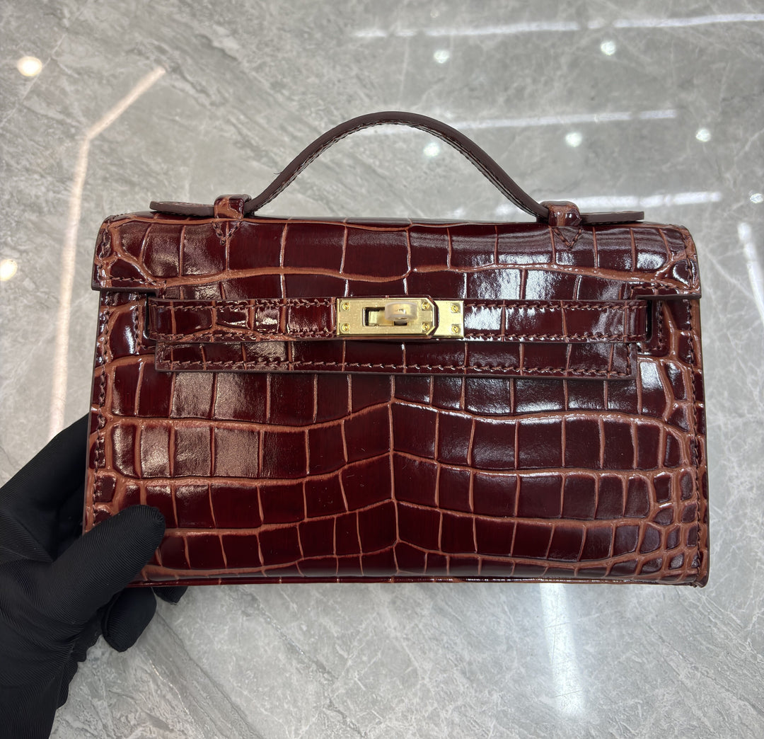 Brown crocodile skin handbag with gold clasp held by a gloved hand on a light gray floor.