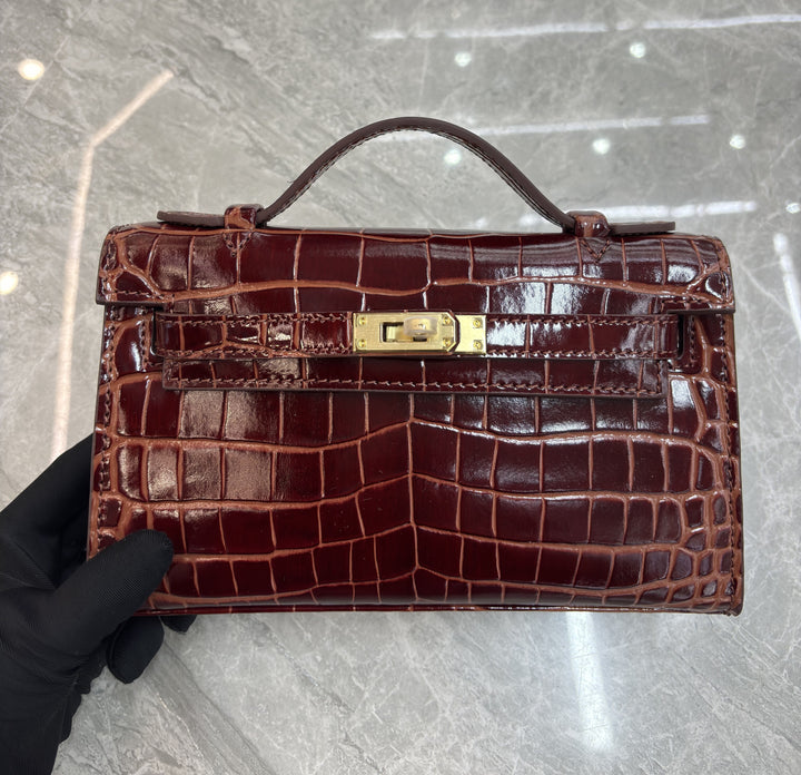 Brown crocodile skin handbag with gold clasp held by a gloved hand on a light gray floor.