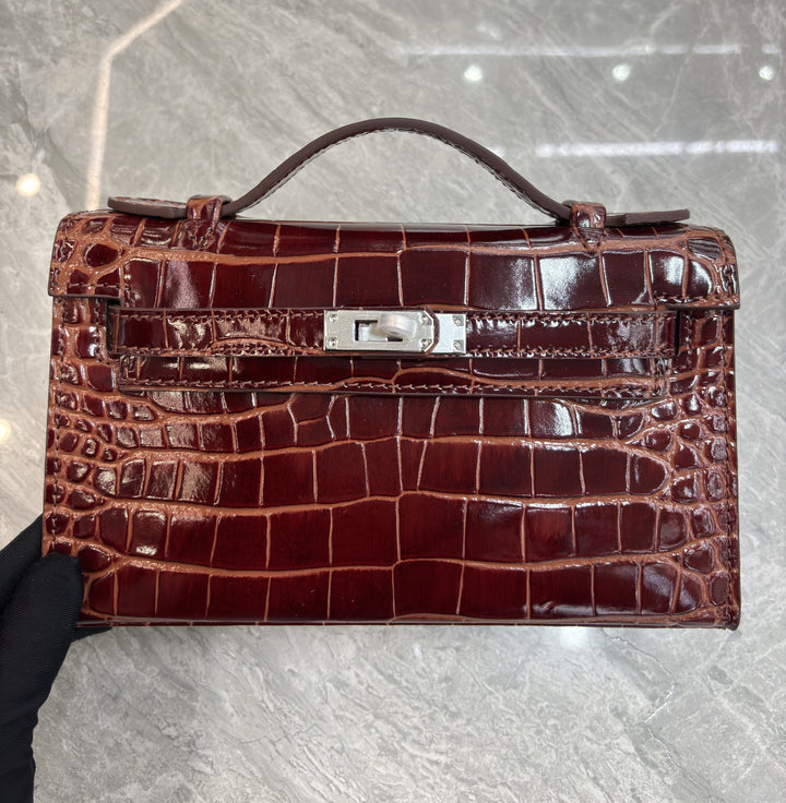 Brown crocodile skin handbag held by a gloved hand on a gray background