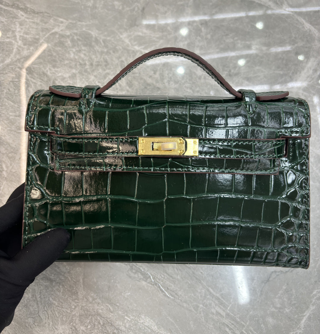 Green crocodile skin handbag with gold clasp held by a gloved hand on a gray background