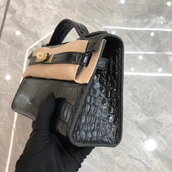 Black crocodile skin handbag with beige interior held by a gloved hand on a reflective surface.