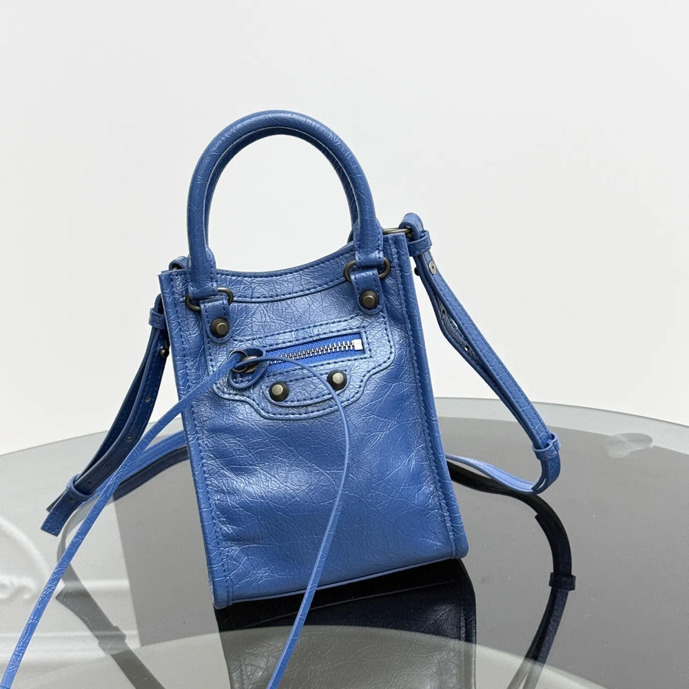 A blue leather top-handle handbag with a detachable shoulder strap, displayed on a reflective glass surface against a light gray background.