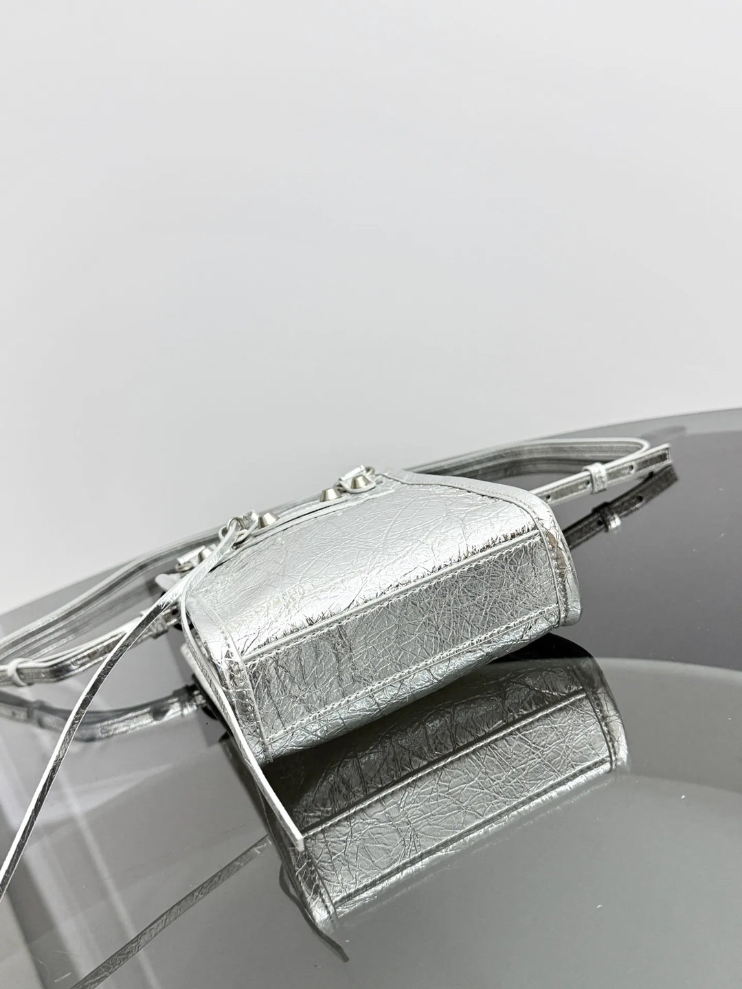 A metallic silver handbag with a textured leather finish, featuring a structured rectangular body, silver-tone metal frame details, and a thin strap, resting on a reflective dark surface against a light gray background.